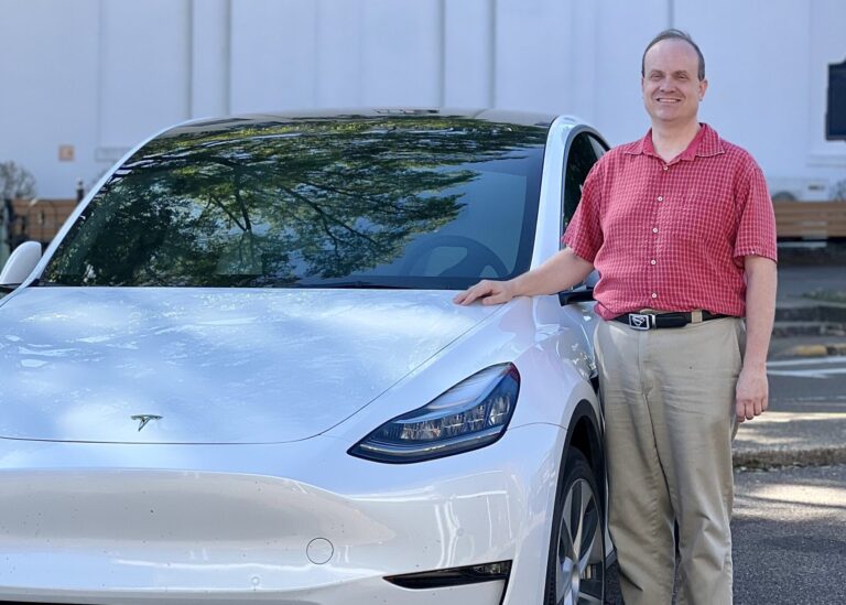 Bill Taylor, dressed in a red shirt and khaki pants, poses alongside his white Tesla Model Y on the Square.