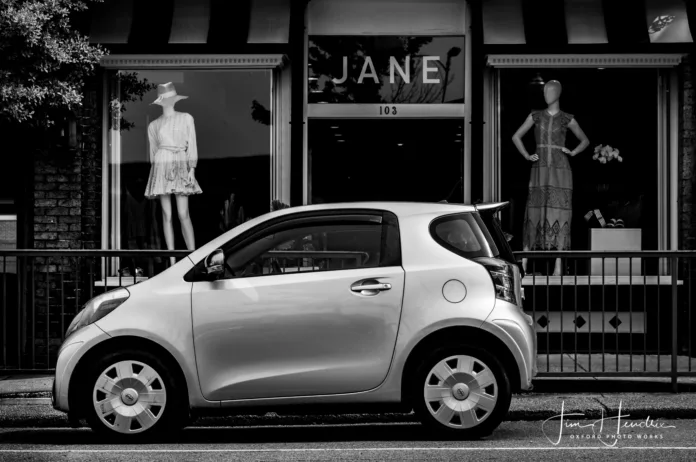 Black-and-white photo of a small modern car parked outside JANE boutique with mannequins in window displays.