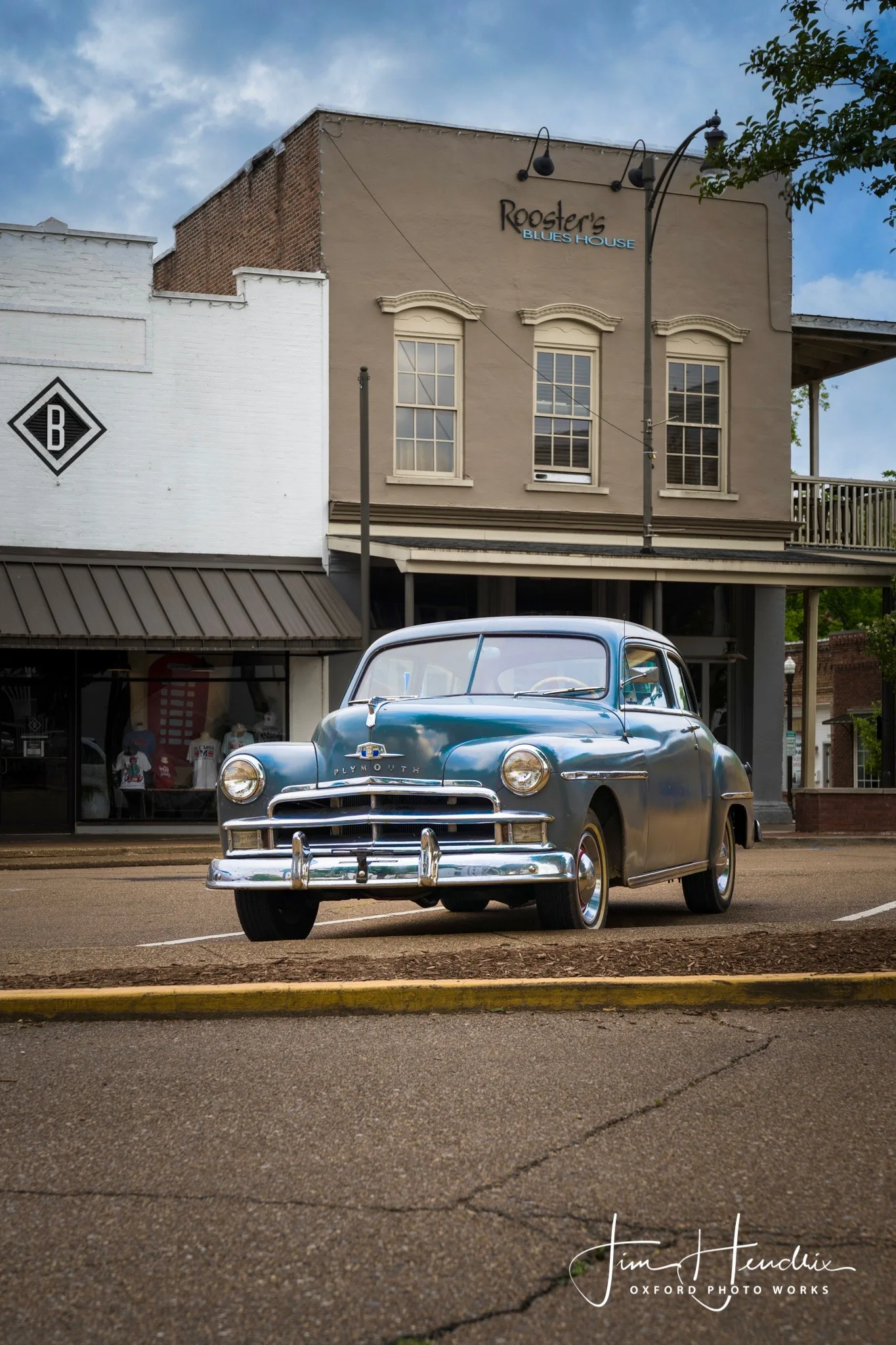 Vintage blue Plymouth parked in front of Rooster’s Blues House on a quiet street under a partly cloudy sky.