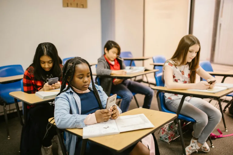 Four students sit at desks in a classroom, each using a smartphone, with notebooks and school materials in front of them.