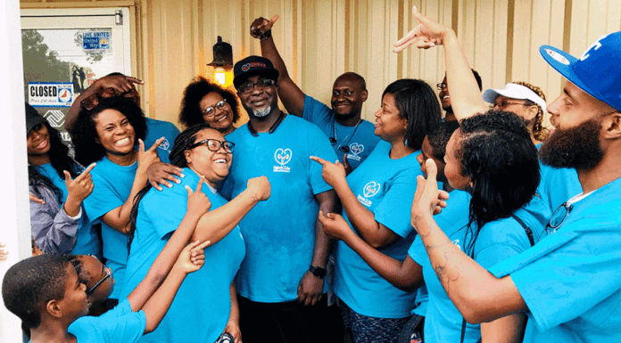 A group of smiling people in matching blue shirts with the Roberta J. Ivy Kidney Awareness Foundation logo gather outside, pointing and celebrating around a man in the center wearing glasses and a cap.