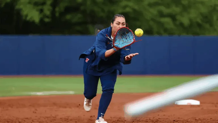 Ole Miss softball player in navy blue uniform reaches forward with her glove to field a yellow softball during a game.