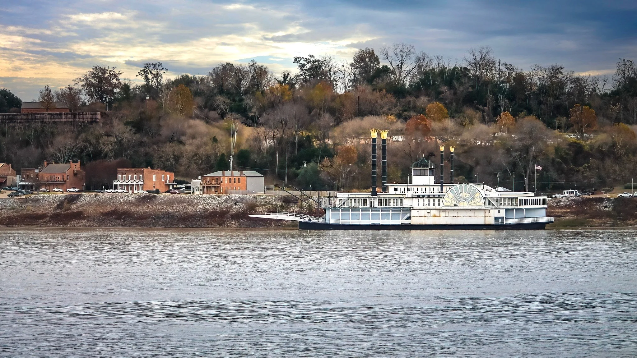White riverboat with twin smokestacks docked by a historic brick town along a calm river under a cloudy sky.