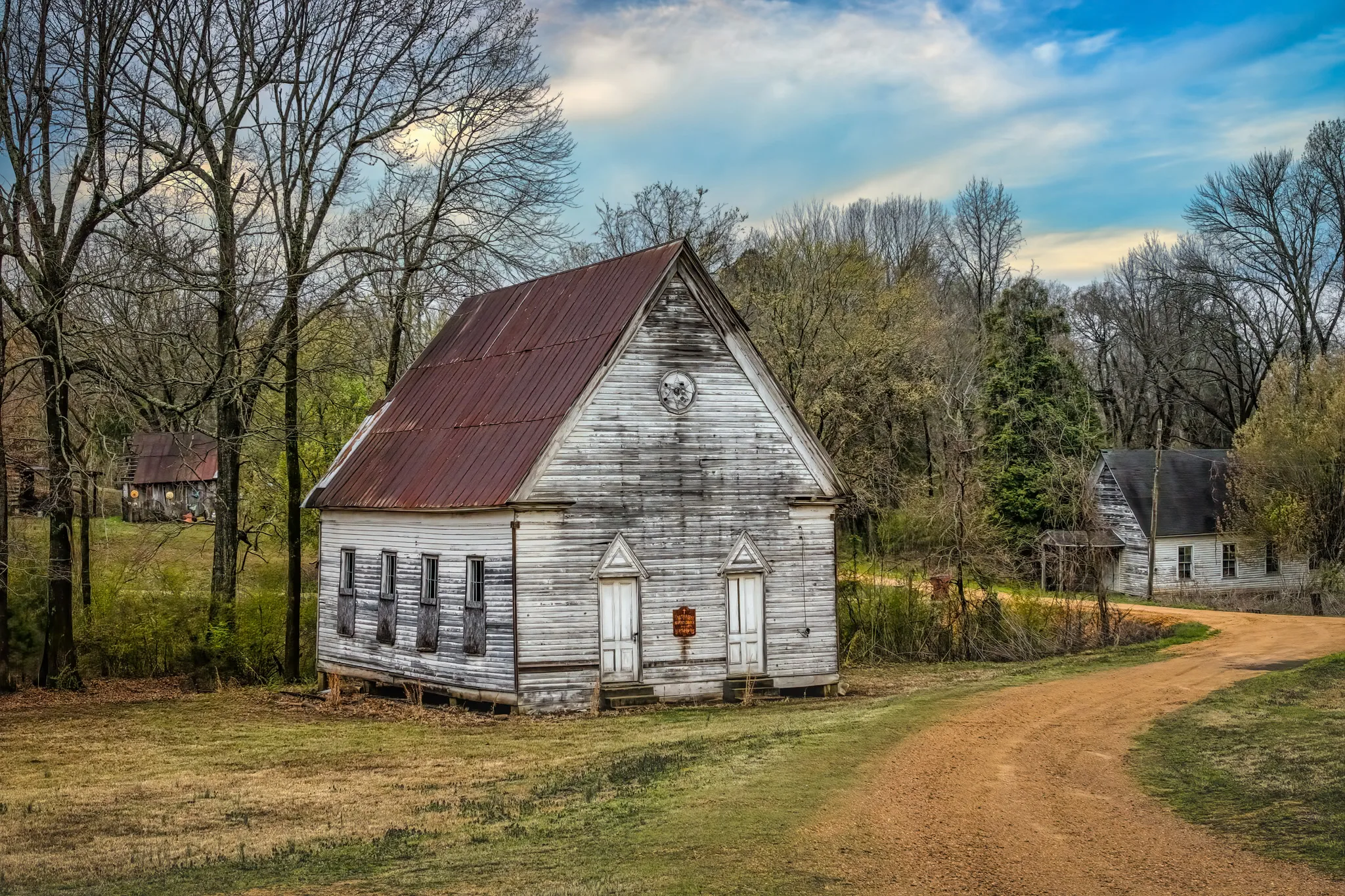 Old wooden church with rusted metal roof beside a winding dirt road in a rural wooded area.