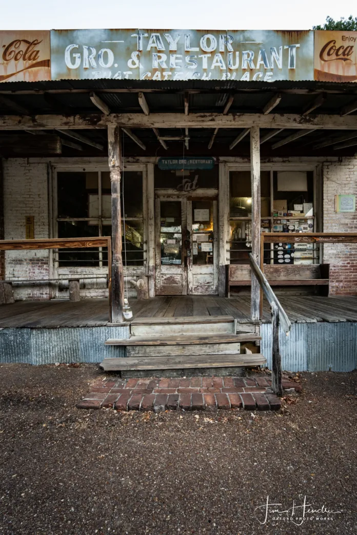 Front porch of Taylor Grocery & Restaurant in Taylor, Mississippi, with weathered wood, faded Coca-Cola signage, and rustic double doors.