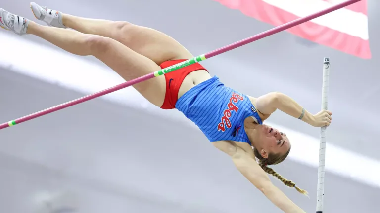 Ole Miss pole vaulter clears the bar during an indoor meet, fully extended mid-air in a blue Rebels uniform with an American flag blurred behind.