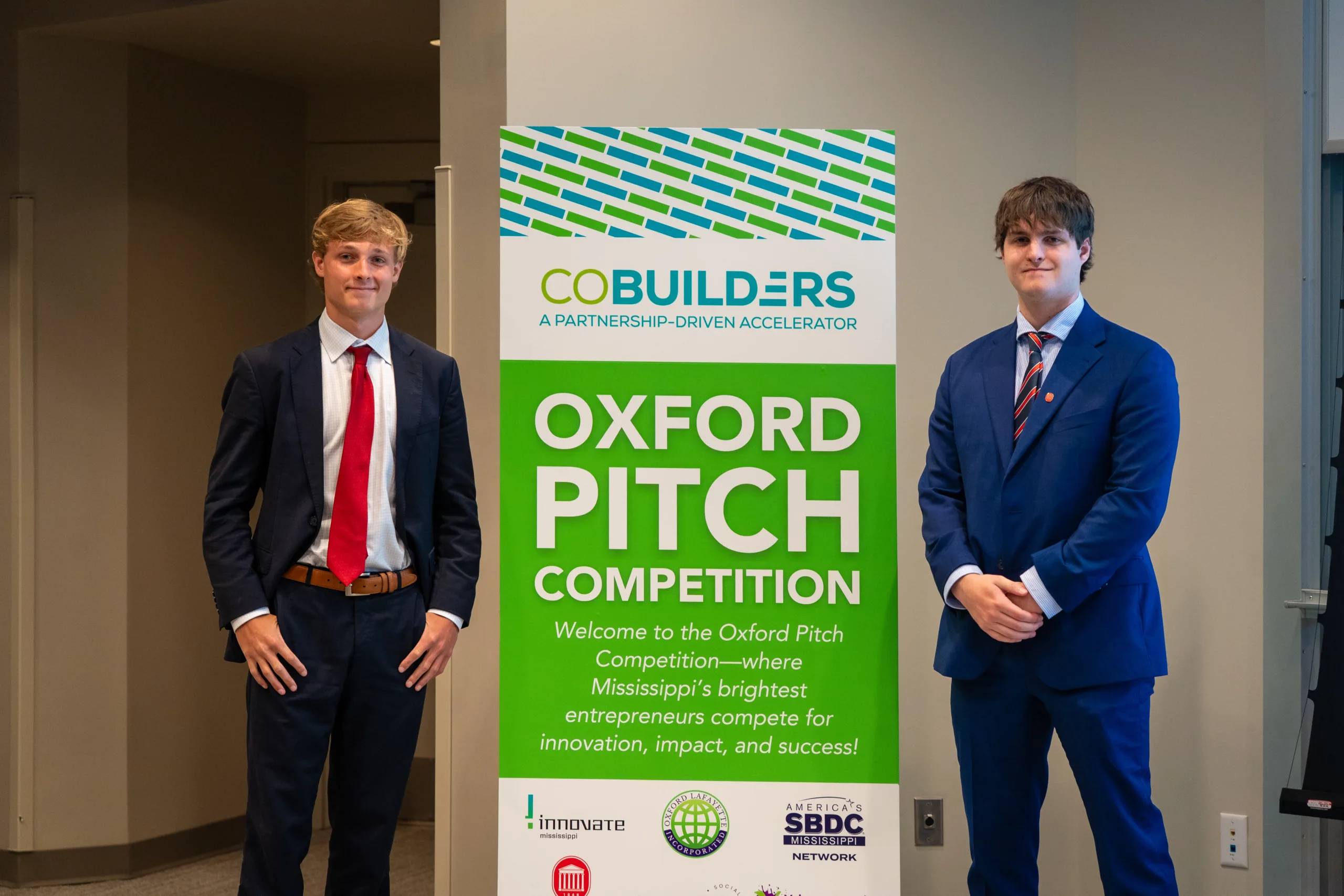 Two Oxford Pitch Competition winners stand beside a CoBuilders event sign.