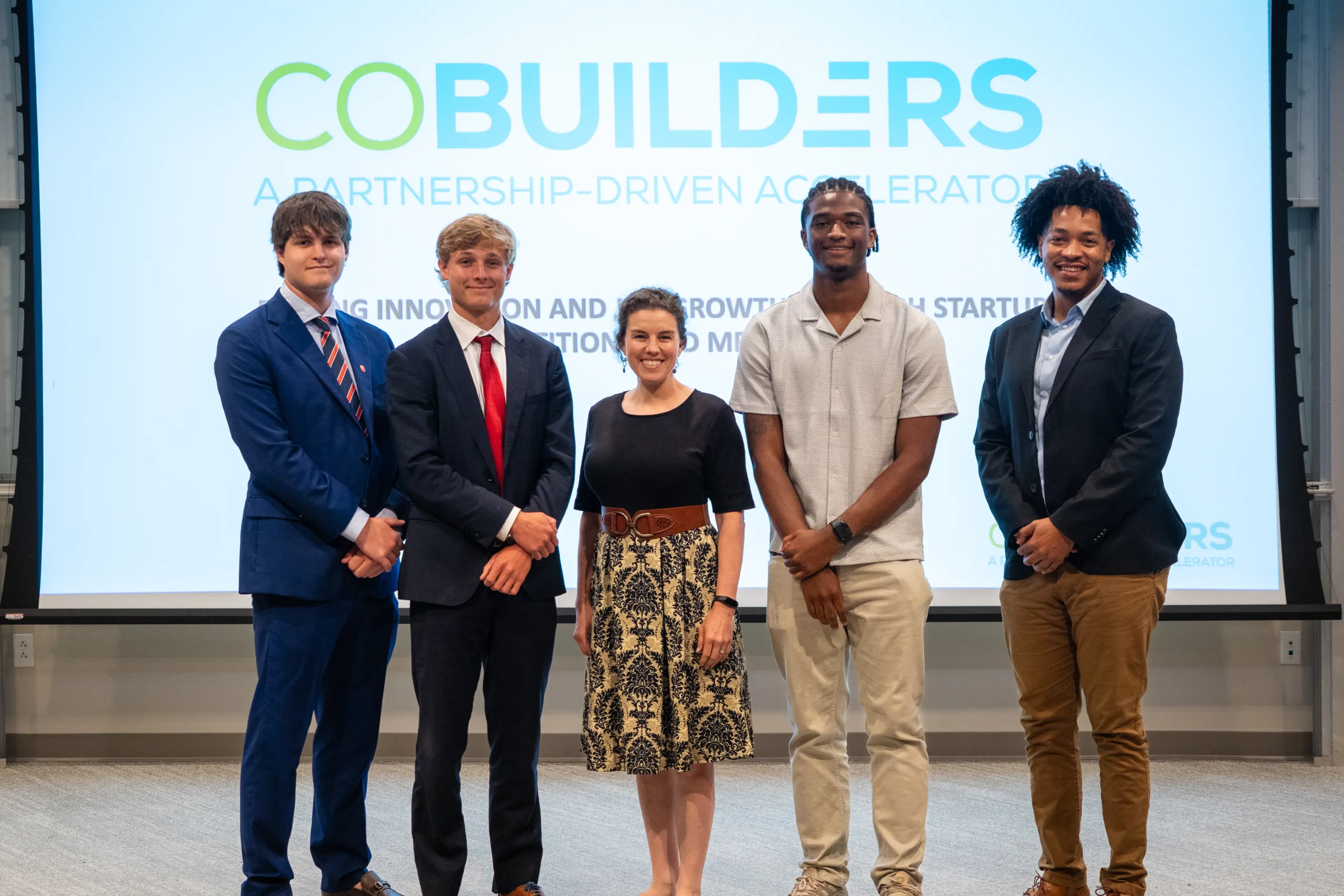 Five Oxford Pitch Competition participants pose in front of a CoBuilders presentation screen.